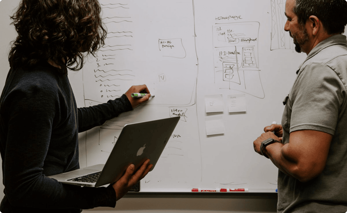 Two people discussing concepts while writing them down on a white board.