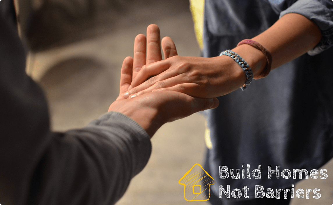To hands clasped together. The text 'Build Homes not Barriers' over the image.