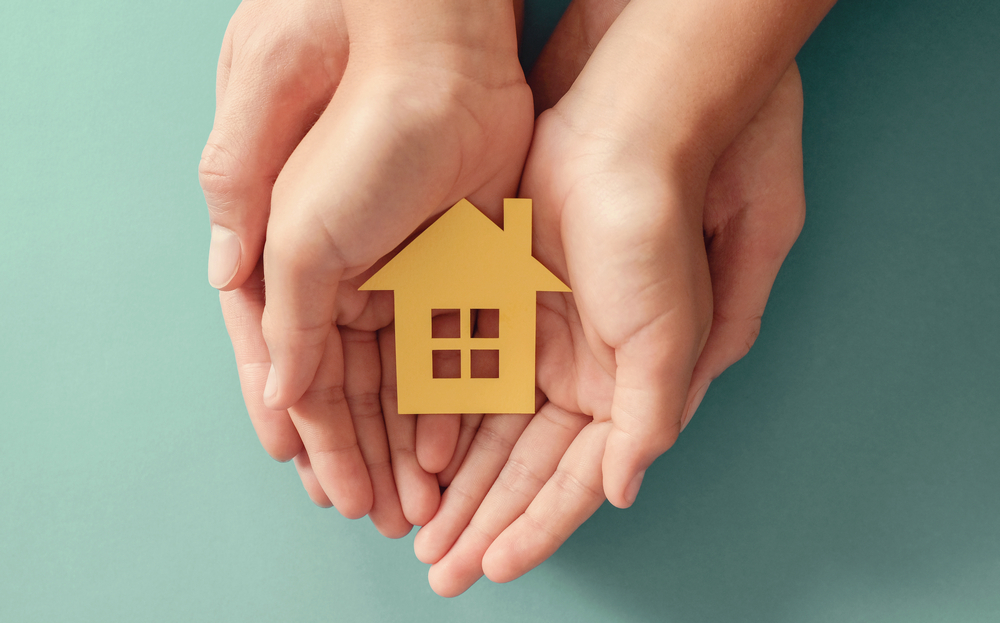 An adult's hands cradling a child's hands cradling a small paper house.